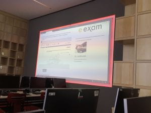 A large projector screen with e-examinations information on it in a room full of computers