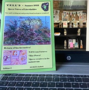 Photo of Summer issue of Tell'd zine - new and exciting art and poetry from local queer creatives - on laptop with photo of Library zine collection on the screen.